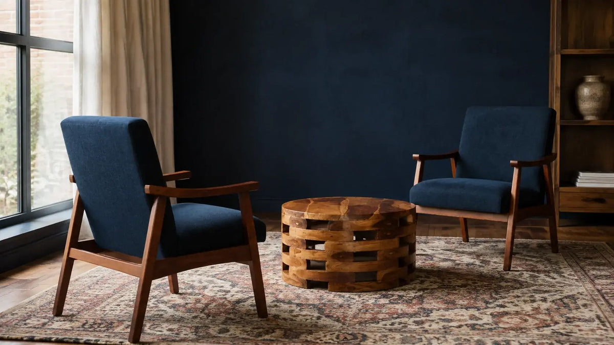 Two navy walnut-frame armchairs face each other across an open-joinery teak drum table on a rust patterned rug in a dark blue room.