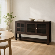 In a room | dark walnut 3-door sideboard against a warm white wall, potted plant at left, soft morning sidelight across the glass doors