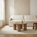 In a room | Round oak-toned coffee table on four pillar legs, on a cream rug in front of a white sofa