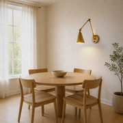 Dining room three-quarter | warm brass swing-arm wall sconce from a different angle above a round pale-oak table, window left, plant right