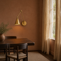 Clay dining room | warm brass swing-arm wall sconce lit on a clay wall beside a dark walnut table, right-side window