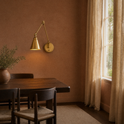 Clay dining room | warm brass swing-arm wall sconce lit on a clay wall beside a dark walnut table, right-side window
