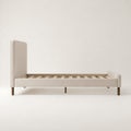 Studio side | Side view of the beige upholstered bed with its headboard, side rail, slat foundation, and dowel legs