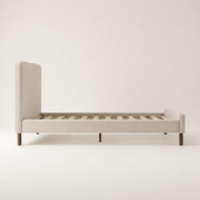 Studio side | Side view of the beige upholstered bed with its headboard, side rail, slat foundation, and dowel legs