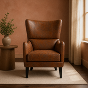 In a clay room | distressed brown embossed faux-leather wingback chair on a plain oatmeal rug, dark walnut floor, round side table and window