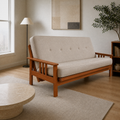 Room | Linen stone futon from a low left three-quarter angle in the cool ivory apartment with pale oak floor and a leafy plant