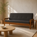 Room | Charcoal-gray futon with honey-toned slatted wood arms in a soft-greige living room with an oak coffee table, lamp, and plant