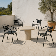 Four black stacking patio armchairs around a round table in a white courtyard beside a potted olive tree
