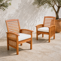 Two warm-brown acacia dining armchairs with ivory seat cushions together on a pale stone terrace