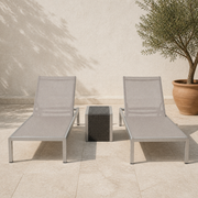 Warm limestone patio | two light gray mesh reclining chaise lounges and dark woven side table beside a potted olive