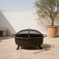 In a room | sunlit limestone terrace with weathered bronze fire pit, seated mesh spark screen and wood-handled poker