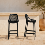 In a room | two black molded-plastic lattice bar stools at a pale terrace bar, olive tree in terracotta pot